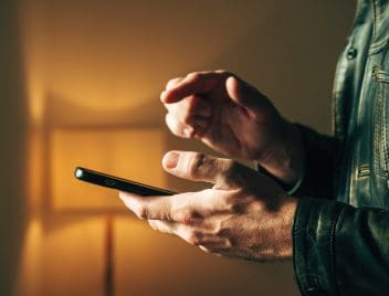 Félhomályban egy bőrdzsekis férfikéz tart egy mobiltelefont, mutatóujjával kattintásra kéeszen.