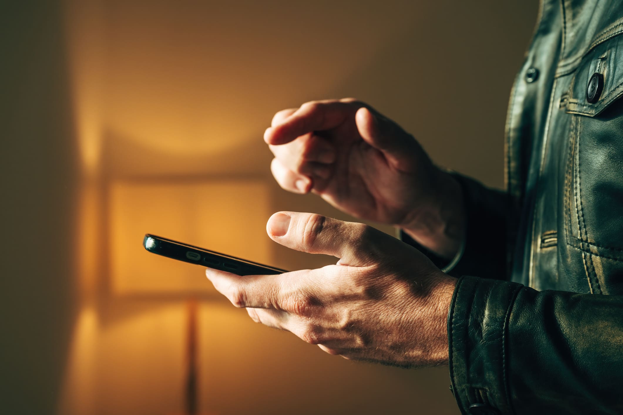 Félhomályban egy bőrdzsekis férfikéz tart egy mobiltelefont, mutatóujjával kattintásra kéeszen.