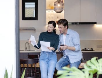 Egy fiatal pár áll az étkezőjüjben egy nyitott teraszajtó előtt, és papírokat nézegetnek. A férfi kezében mobiltelefon.