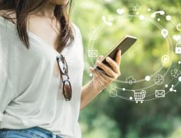 Egy nő kezében látunk egy mobiltelefont, fás háttérrel. A mobultelefon körül egy körívben kis ikonok, amik bármely bankolási tevékenységre utalhatnak.
