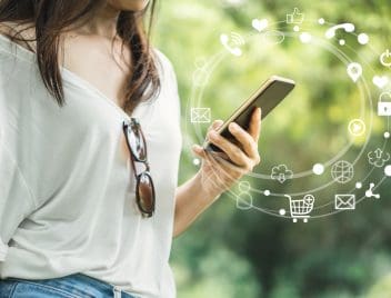 Egy nő kezében látunk egy mobiltelefont, fás háttérrel. A mobultelefon körül egy körívben kis ikonok, amik bármely bankolási tevékenységre utalhatnak.